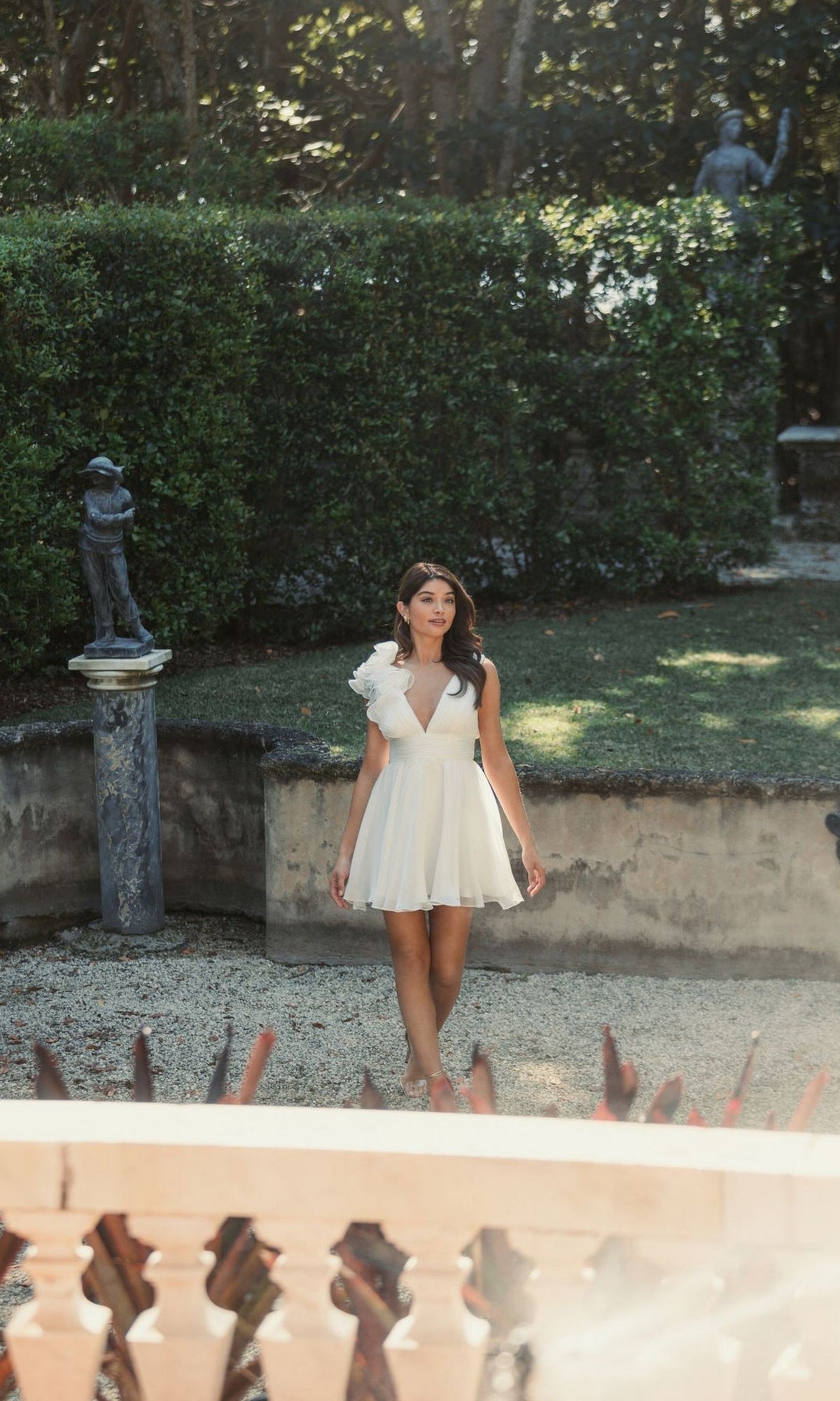 A woman in the Mac Duggal 48952 Ruffle Short White Party Dress strolls through a sunlit garden filled with hedges, statues, and lush greenery.